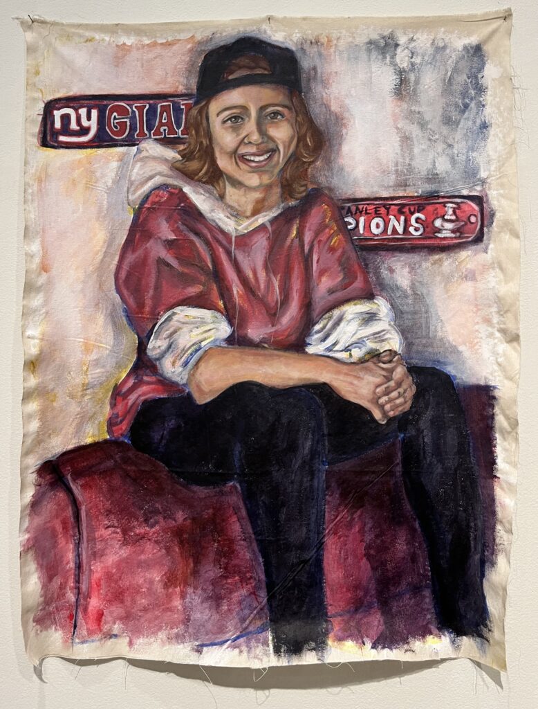 A white person wearing a red and white hoodie, black pants, and a black hat sits on top of a red couch. There is a NY giants and a Washington Capitals stanley cup champion plaque on the wall.