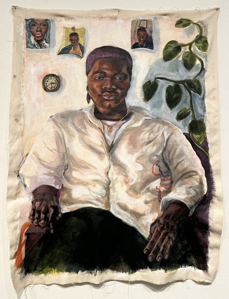 A Black person sits and faces the viewer. He wears a white shirt with a small detail of a red and blue circle on the right side. He is wearing green pants, a white undershirt, a purple earring, and a gold necklace. His hair is dyed purple. There are three photos on the wall of him in drag, his mom, and his best friend. There is also a clock on the wall and a plant coming from the top right.
